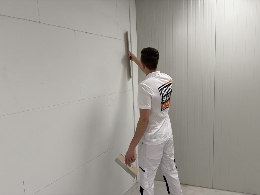Stukadoor staat schuin tegen een lange wand en houdt een rei verticaal tegen de naad, plamuurmes in zijn andere hand